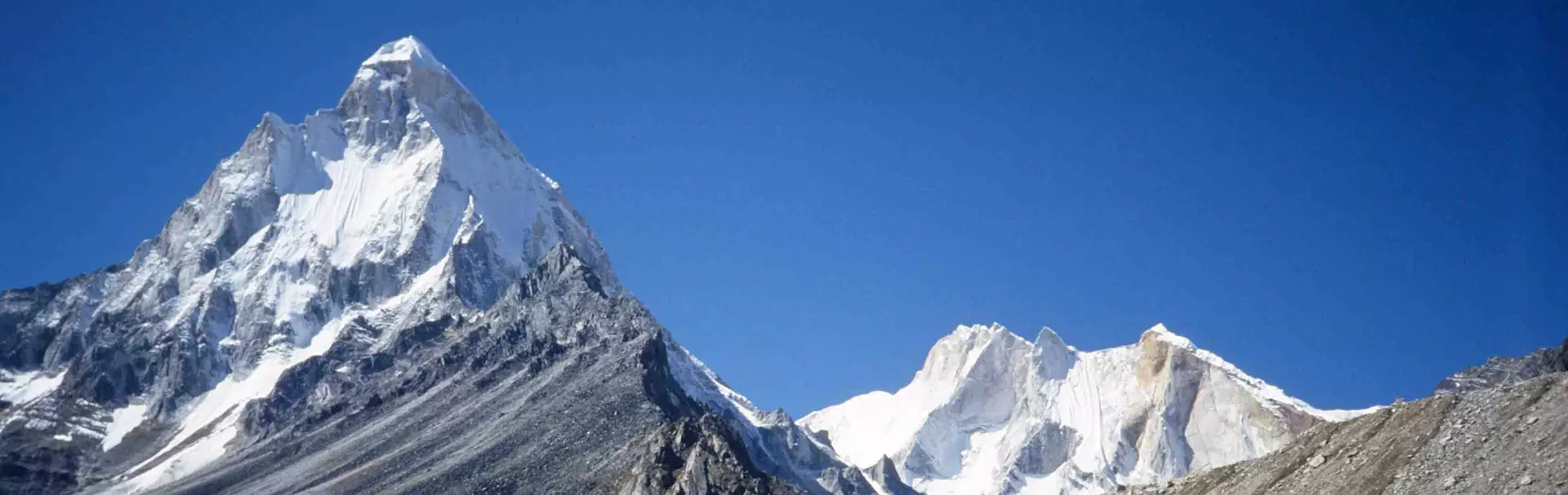 Een bergbeklimmer in rode uitrusting en met een zonnebril zit op de sneeuw op een bergtop, glimlachend met uitgestrekte armen. Op de achtergrond zijn besneeuwde bergtoppen en een helderblauwe lucht te